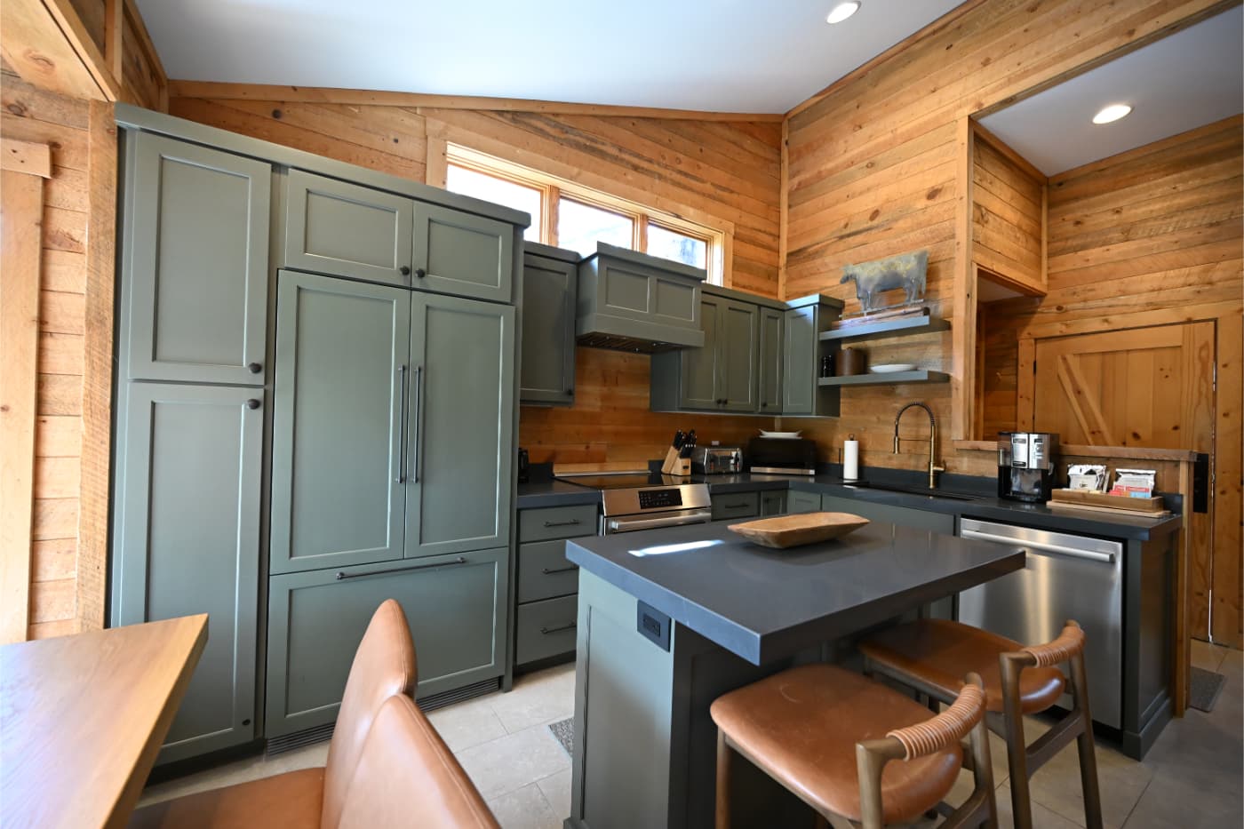 Sage green cabinets and a dark island in a kitchen with knotty pine walls